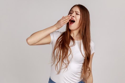 Portrait of attractive tired sleepy woman yawning, closing mouth with hand, exhausted with insomnia, lack of energy, wearing white T-shirt. Indoor studio shot isolated on gray background.