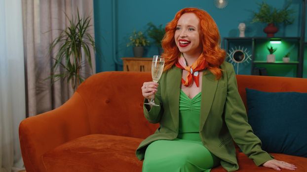 Joyful redhead woman hold glass of champagne cheering and drinking, celebrate success win, career grow up, lucky situation at home in living room. Ginger girl celebrating rent or purchase of new house
