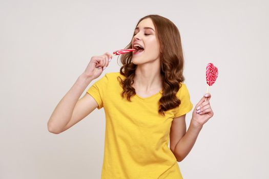 Portrait of woman in yellow casual T-shirt biting lollipop, tasting sweet heart shape candy with satisfied expression, enjoying delicious flavor dessert. Indoor studio shot isolated on gray background