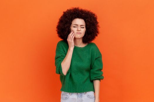 Sick woman with Afro hairstyle wearing green casual style sweater touching her cheek, suffering from tooth ache, frowning face from pain. Indoor studio shot isolated on orange background.