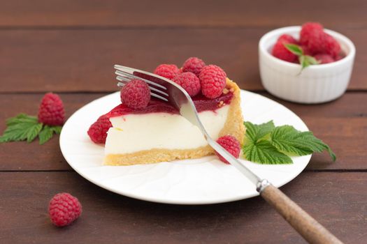 A fork cuts a piece of raspberry pie (cheesecake) lying on a plate, on a wooden background