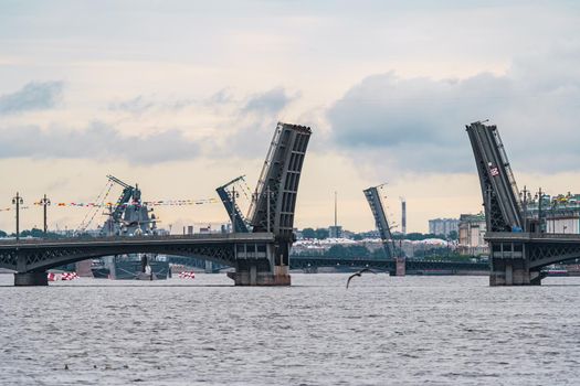 Russia, St. Petersburg, 31 July 2022: A lot of warships goes along the Neva River under open bridges at the celebration of the day of the Navy in sunny weather, drawbridges in background. High quality photo