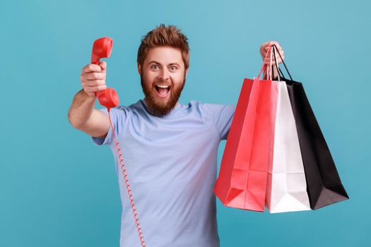 Portrait of excited positive bearded man holding shopping bag and red retro telephone, call me, looking at camera with excitement. Indoor studio shot isolated on blue background.