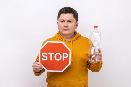 Portrait of serious middle aged man holding plastic bottle and stop red sign, worrying about environment, wearing urban style hoodie. Indoor studio shot isolated on white background.