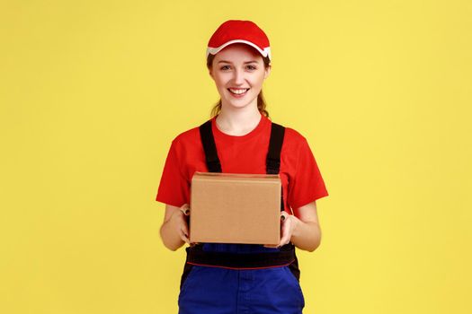 Portrait of delighted courier woman standing with cardboard parcel, giving it to client, looking at camera, wearing overalls and red cap. Indoor studio shot isolated on yellow background.