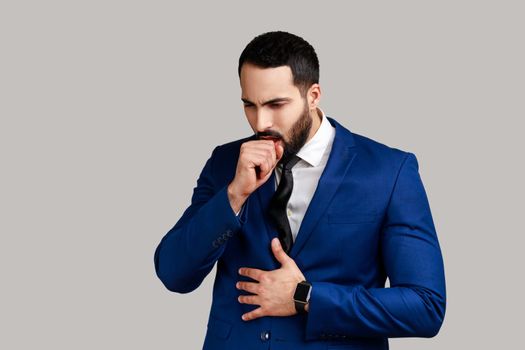 Portrait of unhealthy bearded man coughing, catches cold, having high temperature, having influenza symptom, wearing official style suit. Indoor studio shot isolated on gray background.