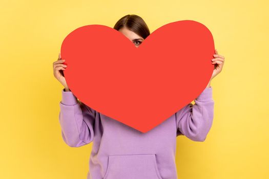 Positive woman hiding her face behind big red heart and looking at camera with curious prying eyes, feeling affection fondness, wearing purple hoodie. Indoor studio shot isolated on yellow background.