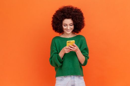 Pleasant looking happy woman with Afro hairstyle wearing green casual style sweater holding smart phone in hands, reading good news in social networks. Indoor studio shot isolated on orange background