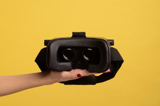 Side view of woman hand holding VR goggles, virtual reality 3d glasses headset for cyberspace simulation, innovative technologies, future and science. Indoor studio shot isolated on yellow background.