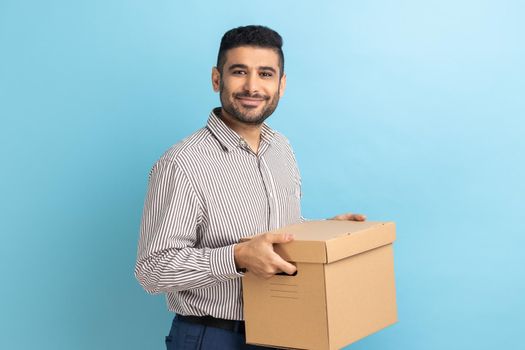 Smiling positive businessman holding cardboard parcel, delivering orders to address, looking at camera with friendly expression, wearing striped shirt. Indoor studio shot isolated on blue background.