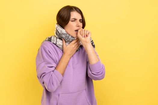 Portrait of unhealthy flu-sick woman standing wrapped in warm scarf shivering from cold, feeling unwell suffering fever, seasonal influenza symptoms. Indoor studio shot isolated on yellow background.