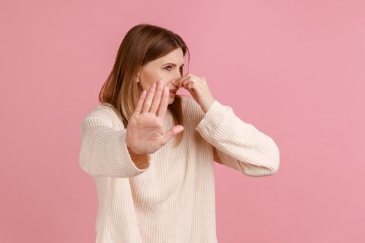 Portrait of woman pinching her nose, holding breath to avoid bad smell and showing stop gesture, disgusted by stinky fart odor, wearing white sweater. Indoor studio shot isolated on pink background.