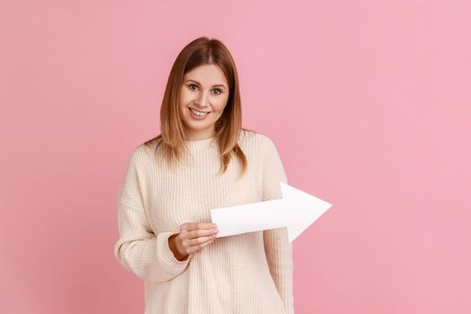 Portrait of smiling satisfied blond woman pointing direction with arrow aside, looking at camera with positive expression, wearing white sweater. Indoor studio shot isolated on pink background.