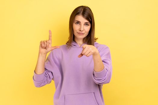 Portrait of frustrated young woman showing looser gesture and pointing at camera, looking with grumpy face, wearing purple hoodie. Indoor studio shot isolated on yellow background.