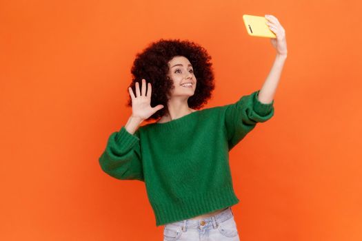 Adorable blogger woman with Afro hairstyle wearing green casual style sweater waving hand to followers while having livestream, greeting new people. Indoor studio shot isolated on orange background.