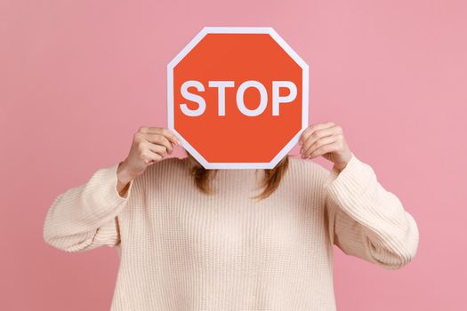 Portrait of unrecognizable woman hiding her face with red sign, campaign against domestic violence, protection of women's rights, wearing white sweater. Indoor studio shot isolated on pink background.