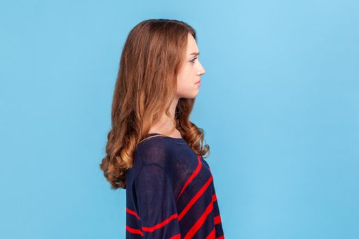 Side view of sad upset young woman wearing striped casual style sweater standing looking ahead, being in bad mood, suffering depression. Indoor studio shot isolated on blue background.