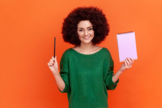 Portrait of happy woman with Afro hairstyle wearing green casual style sweater standing holding paper notebook and pen, looking at camera with smile. Indoor studio shot isolated on orange background.