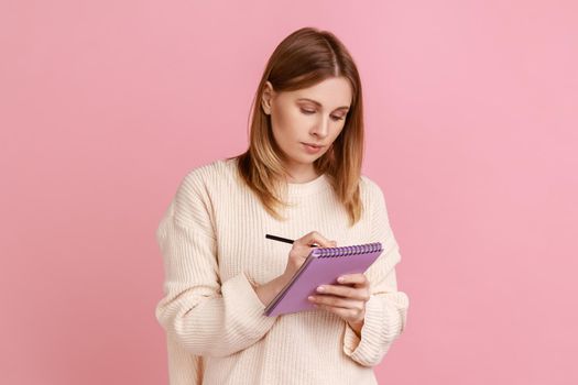 Portrait of serious concentrated blond woman writing in paper notebook, making to do list or list of purchases, wearing white sweater. Indoor studio shot isolated on pink background.