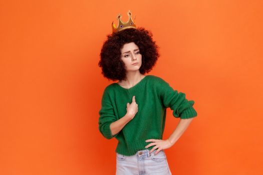 Proud woman with Afro hairstyle wearing green casual style sweater and crown pointing at herself, having narcissistic expression, looking away. Indoor studio shot isolated on orange background.