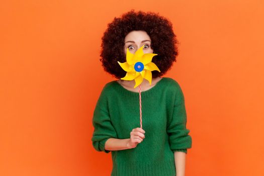 Attractive funny woman with Afro hairstyle wearing green casual style sweater hiding half of her face with yellow windmill, playing, having fun. Indoor studio shot isolated on orange background.