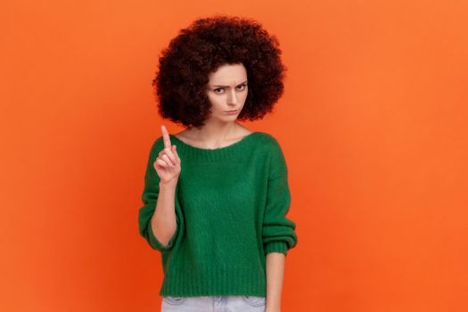 Portrait of serious woman with Afro hairstyle wearing green casual style sweater looking at camera, showing warning attention sign. Indoor studio shot isolated on orange background.
