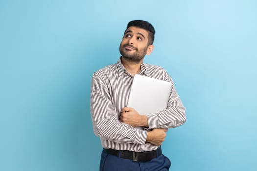 Portrait of pensive businessman standing holding closed laptop or folder, looking away with thoughtful expression, wearing striped shirt. Indoor studio shot isolated on blue background.