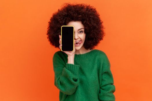Positive funny woman with Afro hairstyle wearing green casual style sweater covering eye with cell phone with blank screen and showing tongue out. Indoor studio shot isolated on orange background.