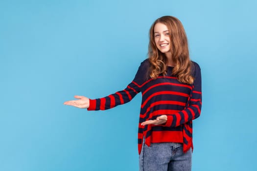 Portrait of smiling woman wearing striped casual style sweater, standing with wide raised arms and welcoming or sharing smth, looking at camera. Indoor studio shot isolated on blue background.