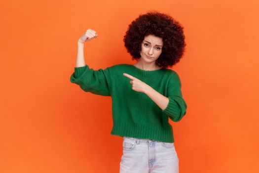 I am strong. Woman with Afro hairstyle wearing green sweater raising hand showing biceps, looking at camera with confidence pride, feeling power. Indoor studio shot isolated on orange background.