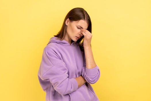 Upset depressed young woman holding her head down, touching face and crying with dramatic grimace, feeling sorrow regret, wearing purple hoodie. Indoor studio shot isolated on yellow background.