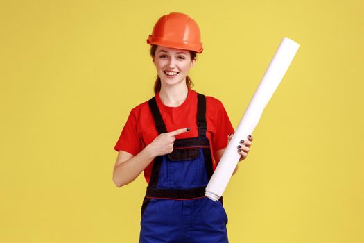 Portrait of satisfied woman architect pointing at blueprints, looking at camera with smile, wearing overalls and protective helmet. Indoor studio shot isolated on yellow background.