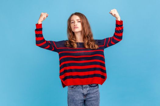 Portrait of confident independent woman, showing biceps, feeling energy, confidence and power in hands to win, become successful, wearing sweater. Indoor studio shot isolated on blue background.