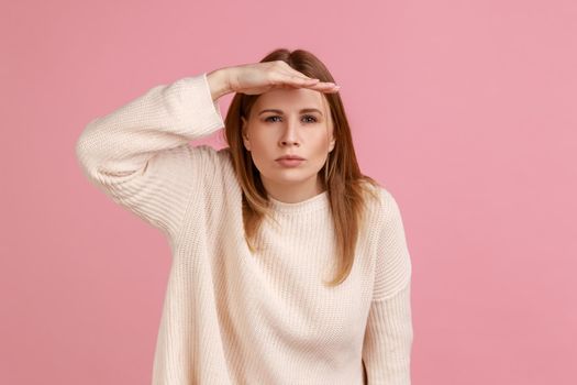 Portrait of blond woman peering into distance, looking far away with attentive view, searching on horizon and curious to discover, wearing white sweater. Indoor studio shot isolated on pink background