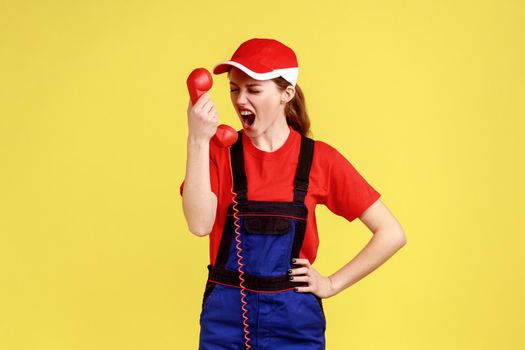 Aggressive worker woman screaming to client on handset, does not accept order, incompetent service personnel, wearing overalls and red cap. Indoor studio shot isolated on yellow background.
