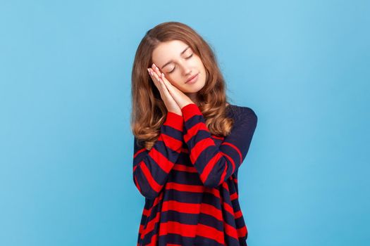 Woman wearing striped casual style sweater, sleeping laying down on her palms and smiling pleased, having comfortable nap and resting, dozing off. Indoor studio shot isolated on blue background.