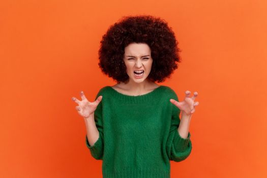 Portrait of angry woman with Afro hairstyle wearing green casual style sweater expressing aggressive emotions, raised arms, screaming. Indoor studio shot isolated on orange background.