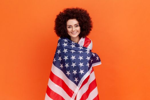 Satisfied woman with Afro hairstyle wearing green casual style sweater standing being wrapped with american flag, expressing positive emotions. Indoor studio shot isolated on orange background.