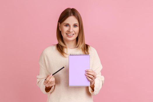 Portrait of happy satisfied blond woman pointing at paper notebook, looking smiling at camera, has positive expression, wearing white sweater. Indoor studio shot isolated on pink background.