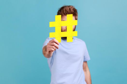 Portrait of bearded man hiding behind yellow hashtag symbol, covering face with hash sign, concept of blogging and viral topics on internet. Indoor studio shot isolated on blue background.