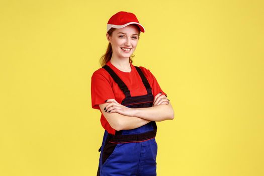 Portrait of adorable worker woman standing with crossed hands, looking at camera with positive confident expression, wearing overalls and red cap. Indoor studio shot isolated on yellow background.