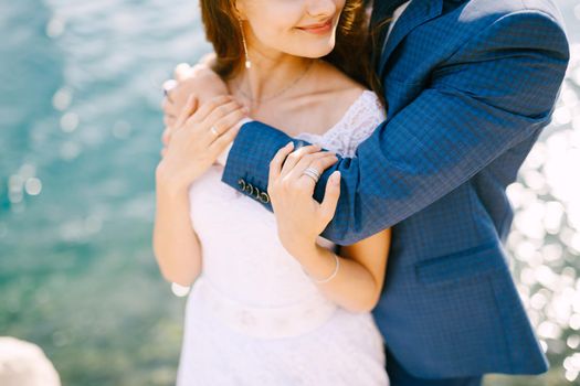 The groom hugs the bride from behind by the shoulders and kisses the forehead on the seashore, close-up. High quality photo
