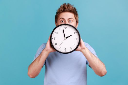 Portrait of astonished handsome young adult man covering half of face big wall clock, time management, schedule and meeting appointment. Indoor studio shot isolated on blue background.