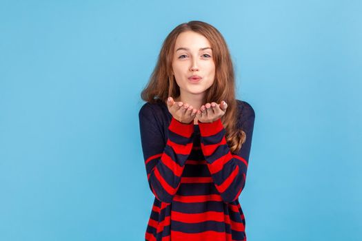 Portrait of romantic woman wearing striped casual style sweater sending air kisses to her beloved man, looking at camera, falling in love. Indoor studio shot isolated on blue background.