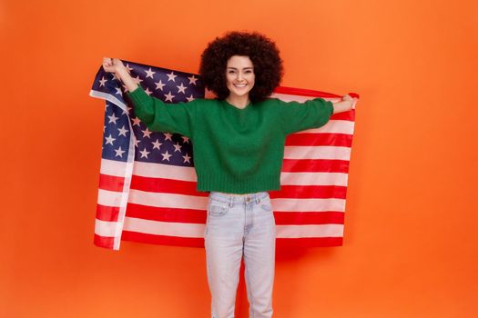 Happy positive woman with Afro hairstyle wearing green casual style sweater standing with raised arms, holding USA flag, celebrating national holiday. Indoor studio shot isolated on orange background.