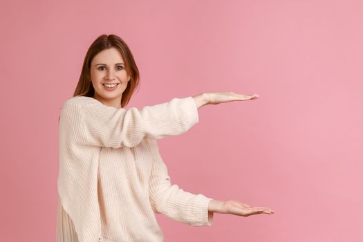 Portrait of happy smiling beautiful blond woman presenting area between hands for advertisement, showing huge size, wearing white sweater. Indoor studio shot isolated on pink background.