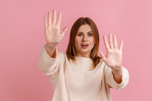 Portrait of scared young adult blond woman gesturing stop with palms and looking surprised with frightened eyes, wearing white sweater. Indoor studio shot isolated on pink background.