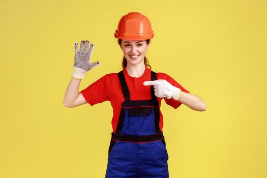 Portrait of woman builder standing with raised arm, pointing at her gloves for protecting hands while working, wears overalls and protective helmet. Indoor studio shot isolated on yellow background