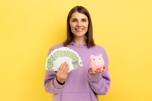 Smiling affirmative woman with dark hair holding big sum of money and piggy bank, profitable investment, wearing purple hoodie. Indoor studio shot isolated on yellow background.