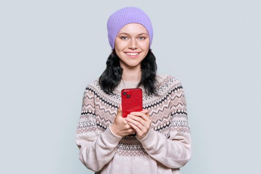 Beautiful young teenage girl in hat in a woolen sweater with smartphone in her hands looking at camera on light gray color studio background. Winter season, Christmas and New Year holidays concept